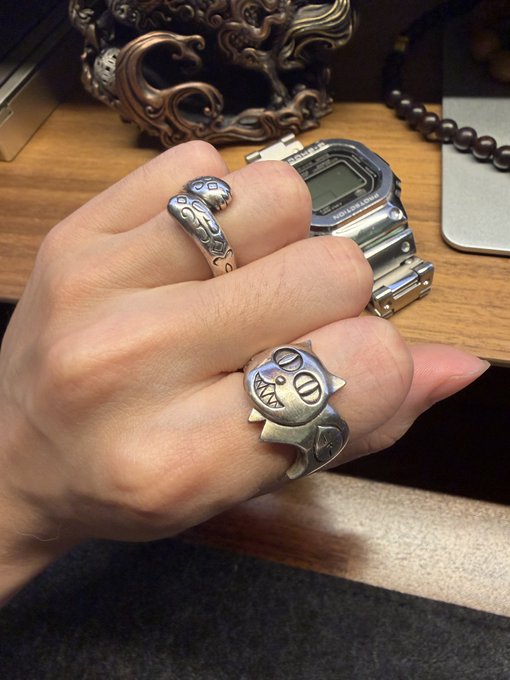 Close-up photo of a hand wearing decorative silver rings, including one shaped like a cat's face with pointed ears and another with raised patterns, alongside a silver digital watch on a wooden surface.