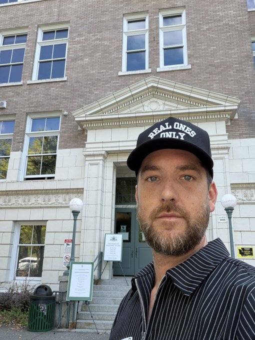 A bearded man wearing a black cap reading 'REAL ONES ONLY' takes a selfie in front of a brick building with classical architectural details.
