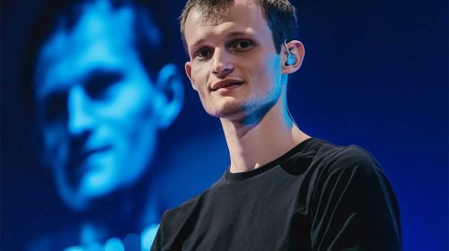 A photograph of Vitalik Buterin, co-founder of Ethereum, speaking at an event with a blue-lit background showing another person's image.