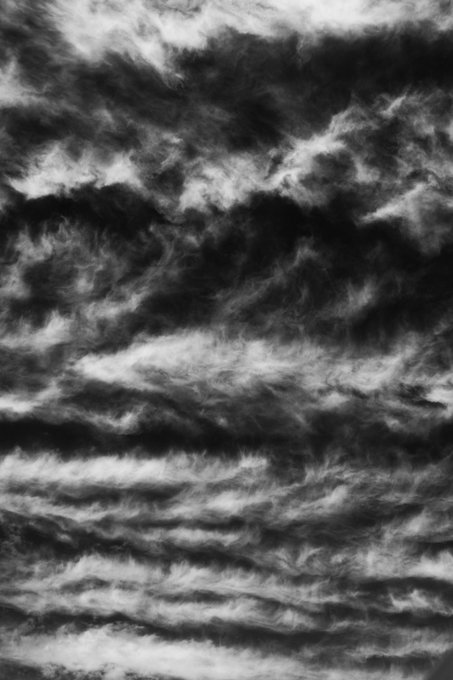 An abstract black and white image showing wispy, hair-like or smoke-like textures flowing horizontally across a dark background.