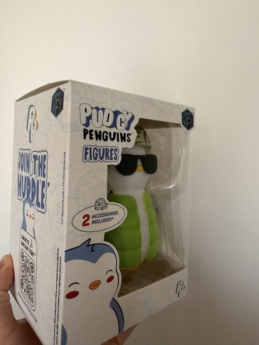 A hand holds a boxed Pudgy Penguins figure wearing sunglasses and a green vest, with packaging indicating "2 Accessories Included" and "Join the Huddle".