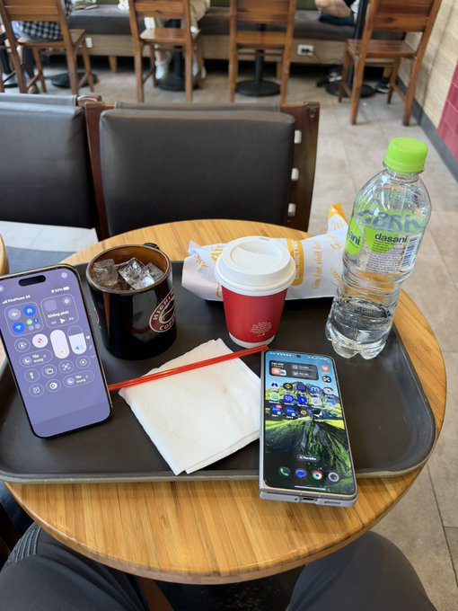 A first-person view of a tray on a wooden table at a cafe or fast food restaurant, containing a smartphone displaying a control center, a Coca-Cola cup with ice, a bottled water, a red coffee cup, a wrapped item, and another smartphone showing a home screen.