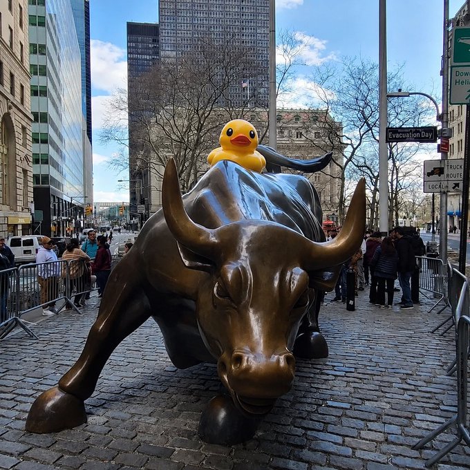 The Charging Bull statue on Wall Street with a large inflatable yellow duck perched on its back, surrounded by tourists and city buildings.
