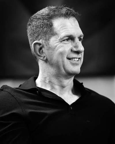 Black and white portrait photograph of a middle-aged man with short graying hair, smiling and looking to the side, wearing a dark collared shirt.