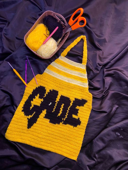 A handmade yellow crocheted tote bag with the word 'GLOVE' stitched in black lettering, displayed on a black fabric background alongside a basket of yellow, black, and cream yarn with orange scissors and a purple crochet hook.