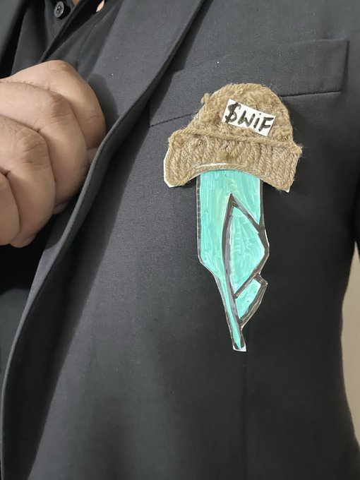 A close-up of a hand-drawn "Knife" badge pinned to a black suit jacket, depicting a turquoise blade topped with a brown fuzzy hat.