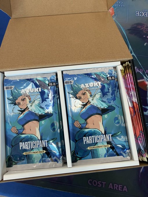 A cardboard shipping box labeled "COST AREA" contains multiple sealed "Azuki Tournament Card Game" Participant Tournament Packs (Ages 13+, Vol. 01) featuring blue-haired anime character artwork, supporting the post's praise of Azuki's TCG direction.