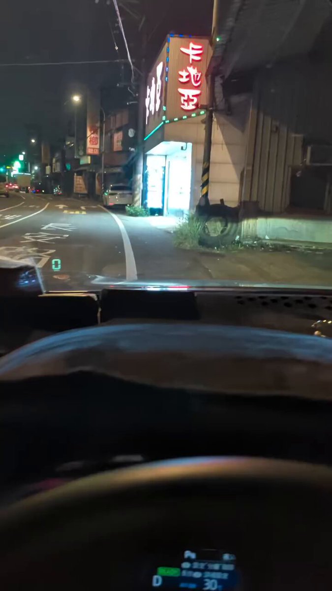 The image shows a view of an interior from inside a car, with the focus on the dashboard display screen. The windshield wipers are on, indicating that the car is active. A street light can be seen near a business building at night. There is also text in the image, reading "台灣檳榔西施你們看過嗎", which seems to have been edited or altered. The focus of the photo appears to be on the car's interior and its surroundings rather than the posted content in the image itself.
