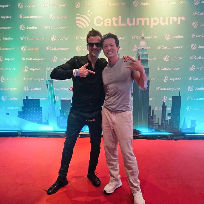 Two men pose cheerfully in front of a "CatLumpurr" backdrop featuring repeated "Jupiter" and "CatLumpurr" logos alongside a city skyline, with the man in the black jacket giving a thumbs-up—capturing the excitement of a past event that the caption eagerly anticipates will return soon.