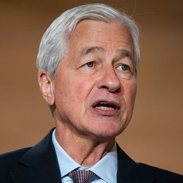 A close-up portrait of an older man with gray hair, wearing a dark suit and light blue shirt, looking off to the side as if speaking.
