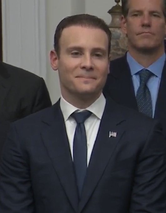 A photo of a man in a dark suit and blue tie with an American flag pin, standing in what appears to be the White House.