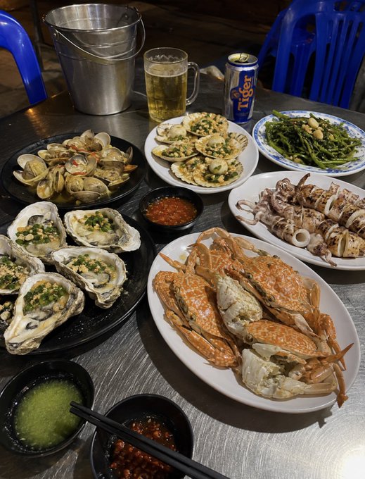 A table spread featuring a variety of seafood including clams, scallops, oysters, squid, and large crabs, accompanied by a mug of beer, a can of Tiger beer, and dipping sauces.