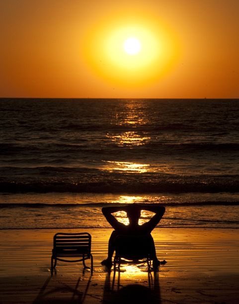 A silhouetted figure relaxing in a beach chair at sunset, with the golden sun reflecting across the ocean waves and an empty chair beside them.