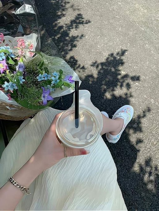 A woman in a flowy cream dress holds an iced coffee with a black straw alongside a bouquet of wildflowers, wearing white sandals and a gold chain bracelet on a sunlit street. The image captures a peaceful, aesthetic "good morning" moment that pairs with the post's GM (good morning) greeting and Bitcoin-focused mindset, suggesting a calm, productive start to the day.