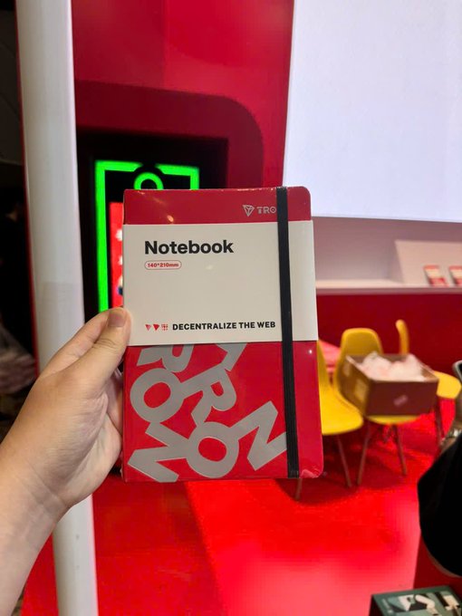 A hand holds up a red TRON-branded notebook with the text 'Notebook', '140*210mm', and '♥♥ DECENTRALIZE THE WEB' on its cover, taken at what appears to be a TRON event booth.