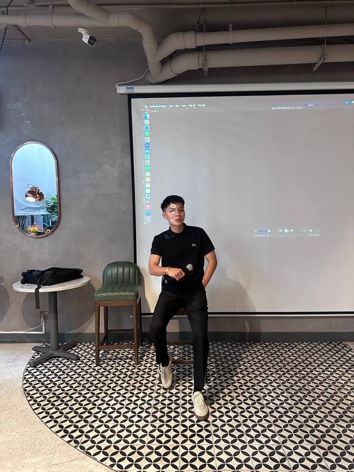 A young man in a black polo shirt and pants sits on a green stool in a modern room with a projector screen, round mirror, and patterned tile floor.