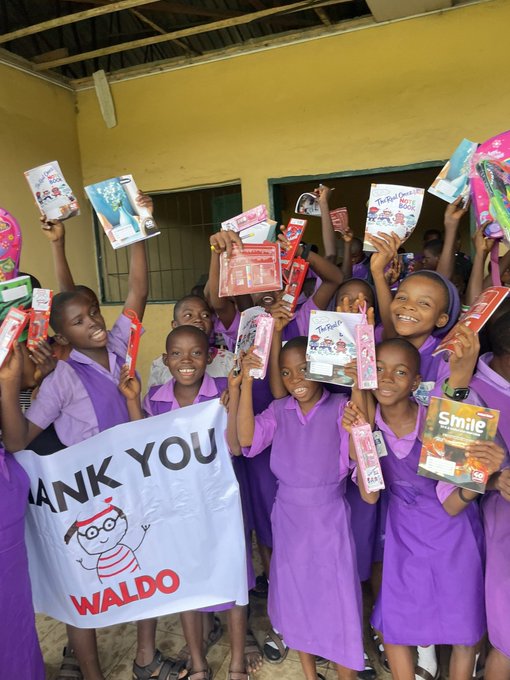 Group of smiling school children in purple uniforms holding up school supplies and a white banner that says 'THANK YOU WALDO' with the Waldo Books logo.