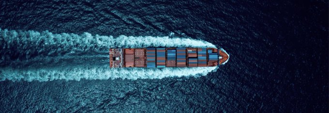 An aerial view of a cargo ship loaded with containers cutting through dark blue ocean water, leaving a white wake.