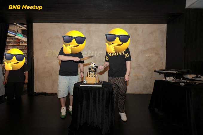 Three people with their faces covered by sunglasses emojis stand at a "BNB Meetup" event around a small black table with a skull figurine and a cake, with "BNB" visible on the wall behind them.