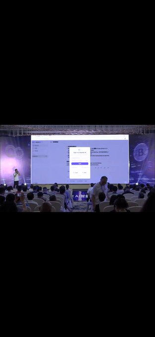 A conference or presentation is taking place with a speaker on stage in front of a large screen displaying a sign-up form with fields for "First Name," "Email," and a "Sign Up" button, while an audience sits in chairs watching. A Bitcoin (₿) logo is visible on the right side of the stage backdrop.