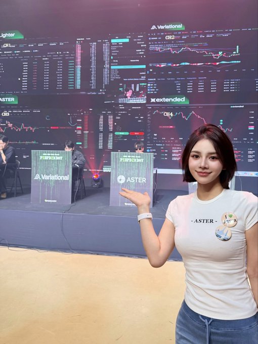 A woman wearing a white Aster-branded t-shirt gestures toward a stage where a Variational and Aster-sponsored cryptocurrency trading competition is displayed on large screens showing market data.