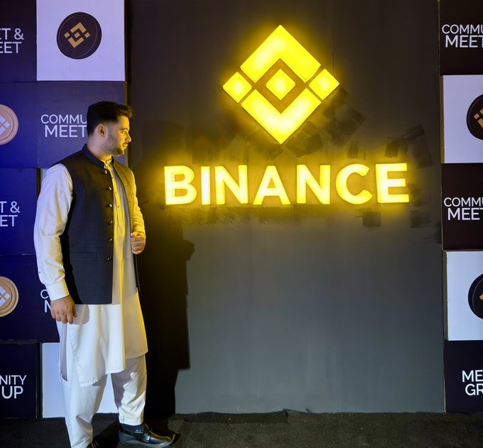 A man dressed in traditional attire (white shalwar kameez and a dark vest) standing in front of an illuminated yellow 'BINANCE' sign at what appears to be a community meet event.