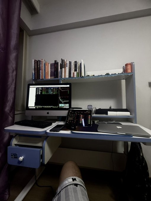 A home desk setup with an iMac displaying stock charts, a tablet, keyboard, and books on an overhead shelf above a blue drawer unit, with a person's leg visible underneath.