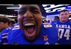 A young man in a blue Kansas jersey shouts excitedly into the camera at a sporting event, with another fan wearing a #74 jersey visible behind him.