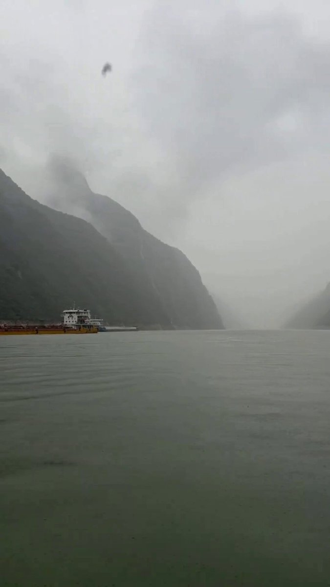 The image shows a boat floating on a body of water, possibly a lake or river. The sky above appears cloudy, creating an overcast atmosphere. There are two boats visible in the scene - one near the center of the image and another towards the right side. A bird can be seen flying high up in the sky, adding to the overall ambiance of the scene.