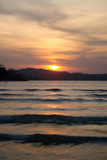 A serene sunset over a calm body of water, with the orange sun dipping below a distant, silhouetted shoreline beneath soft, wispy clouds.