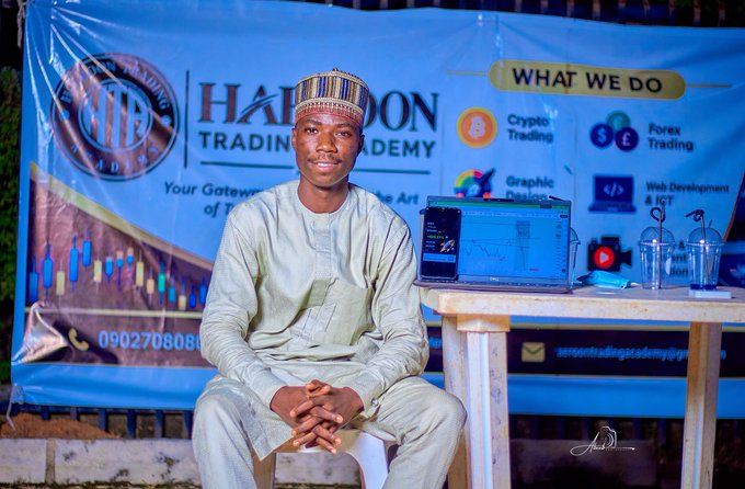 A young man wearing traditional attire and a cap, seated in front of a 'Halibon Trading Academy' banner advertising services in crypto, forex, graphic design, and web development.