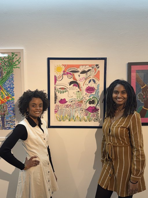 Two smiling women stand in an art gallery in front of colorful framed paintings, one wearing a white pinafore dress and the other a brown striped dress.