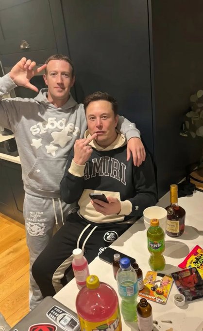 Two men pose playfully behind a cluttered table covered with empty bottles, soda cans, and cigarette packs, with one making a frame gesture and the other holding a cigarette while checking his phone. The man on the right wears an "AMIRI" hoodie and appears to be Elon Musk, connecting the post's @elonmusk tag to a casual, candid moment with a companion amid an evening of drinks and snacks.
