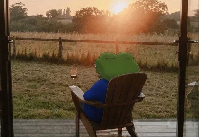 A plush toy resembling a frog wearing a blue shirt sits in a wooden Adirondack chair on a deck, watching a sunset over a grassy field.