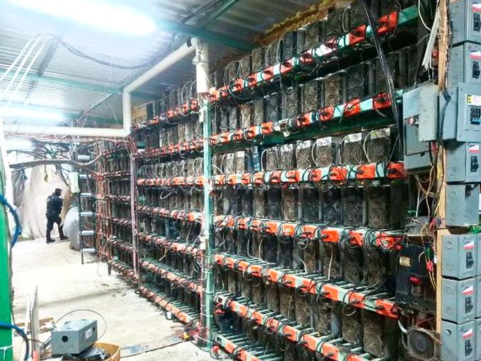 A large warehouse filled with rows of cryptocurrency mining rigs stacked on metal shelving, with extensive wiring and electrical panels visible throughout the facility.