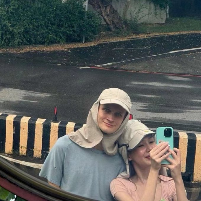 In the image, there are two people posing for a picture next to each other while one of them holds a cell phone with their face visible. The couple is standing near a fence or railings that line the street. In the background, a car can be seen parked on the side of the road. Above all this, it appears that they have attracted the attention of an Instagram user who has posted their picture and captioned it: "Vitalik got a girlfriend Ethereum to $10,000 now". The focus in the image is primarily on the couple posing for a photo while the social media post captures their moment together.