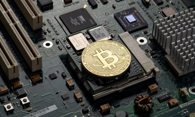 A gold Bitcoin token rests on a computer motherboard, symbolizing the intersection of cryptocurrency and hardware technology. The image ties into the post's theme about China's recurring threats to ban Bitcoin, visually highlighting the digital asset at the center of these regulatory debates.