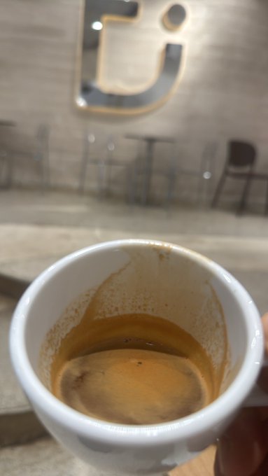A photograph of a hand holding a white ceramic mug containing the last remnants of an espresso shot in a cafe setting, with a stylized metal logo on the wall in the background.