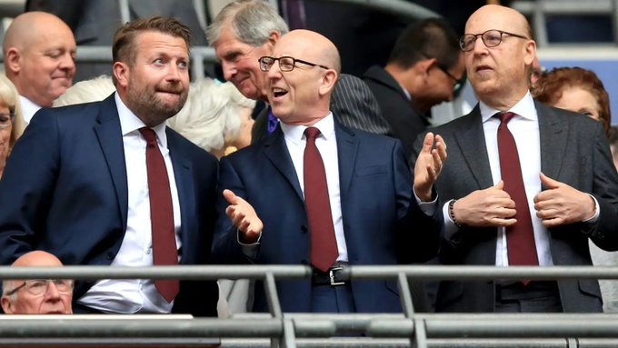 Three men in dark suits with red and blue ties watching from stadium seating, with the central man gesturing animatedly with his hands.