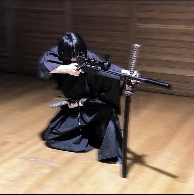 A person dressed in dark traditional samurai robes kneels on a wooden floor, aiming a modern sniper rifle through a scope while a katana rests beside them, blending ancient and modern weaponry. The image pairs with the post's "Ready to attack the day?" message, symbolizing a warrior mindset and readiness to take aim at goals with precision and intensity.
