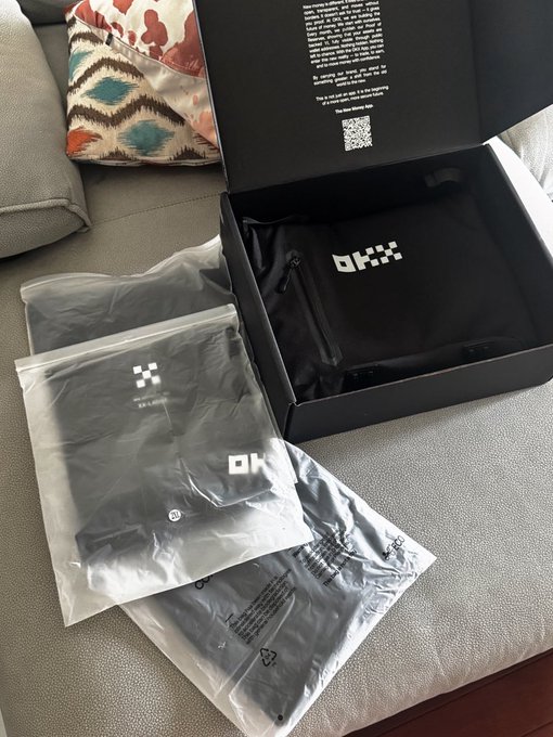 A photo of an 'OKX' branded merchandise unboxing, showing black clothing items in packaging on a couch with a patterned pillow.