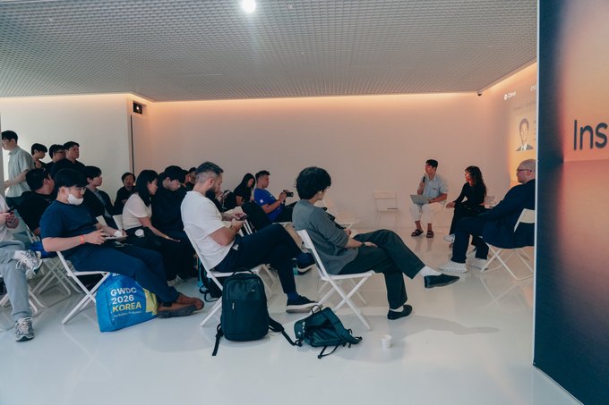 An audience attends a panel discussion at an 'Inst' event, with speakers seated on white chairs against a cream wall.