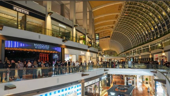 An interior view of a multi-level shopping mall with a curved glass ceiling, featuring storefronts including 'MARQUES SPORTSCLUB' and 'PARTY' on the upper level, with crowds of people gathered along the railings.