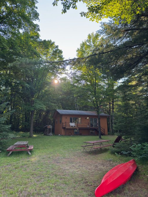 A wooden cabin surrounded by tall trees at sunset, with a picnic table, grill, and a red kayak resting on the grass in the foreground.