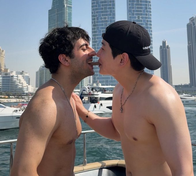 Two shirtless young men playfully interacting with their mouths open toward each other on a boat, with a city skyline of high-rise buildings visible in the background.