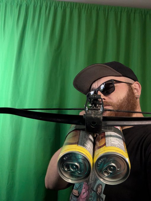 A bearded person wearing a cap and sunglasses holding a makeshift crossbow made from two beverage cans taped together, set against a green screen backdrop.