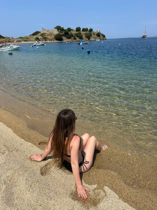 A woman in a bikini sits on a sandy beach with crystal-clear water, with a small fortress-topped island and boats visible in the background.