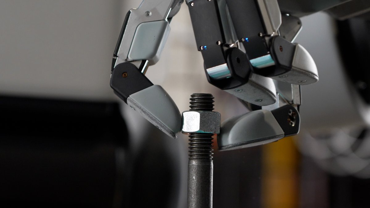 A close-up shot shows a robotic hand gripping a metal bolt and nut on a threaded rod.