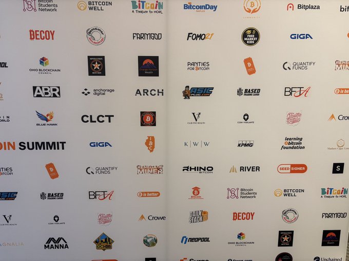 A backdrop wall features numerous cryptocurrency company logos, including Bitcoin Well, BitcoinDay, and Bitplaza, associated with a Bitcoin Summit.