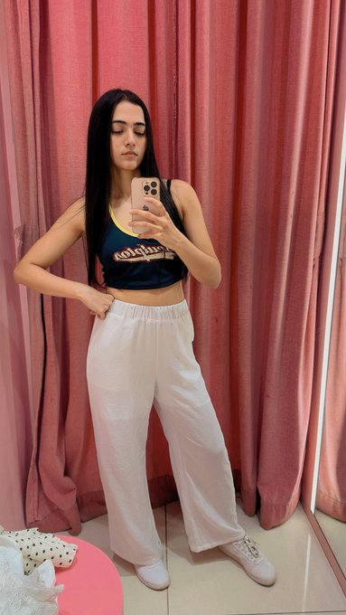 A young woman takes a mirror selfie wearing a dark crop top with "Colgate" branding and white wide-leg pants, standing against pink curtains. The image appears to showcase her new outfit, likely from the shopping trip mentioned in her post about getting back to her "CT duties."